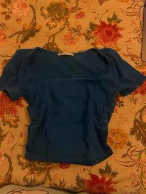 Blå topp med rynkade sidor från Zara - Säljer en blå topp från Zara i storlek S. Toppen har korta ärmar, fyrkantig halsringning och snygga rynkade detaljer längs sidorna. Materialet känns mjukt och stretchigt, perfekt för dig som gillar en enkel men stilren look.