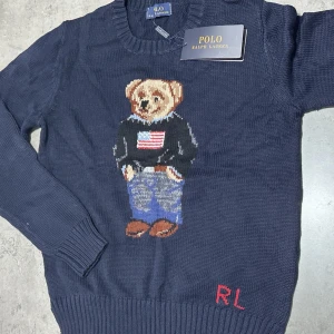 Mörkblå stickad tröja Polo Ralph Lauren - Säljer en mörkblå stickad tröja från Polo Ralph Lauren med ikonisk björn på framsidan. Tröjan har rund hals, långa ärmar och ribbade muddar. RL-broderi i rött nedtill. Perfekt för dig som gillar klassisk och cool stil.