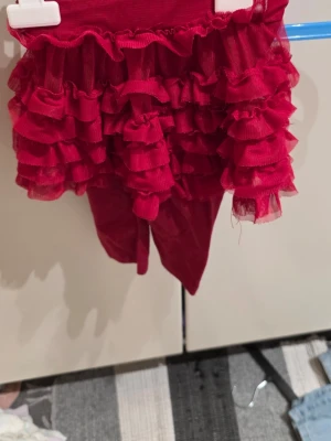 Röda leggings med volangkjol från Mywearbaby - Säljer ett par röda leggings med fastsydd volangkjol i mesh från Mywearbaby, storlek 68. Kjolen har flera lager volanger och leggingsen har ett broderat hjärta på benet. Kjolen är i polyester och leggingsen i bomull med lite elastan för stretch.
