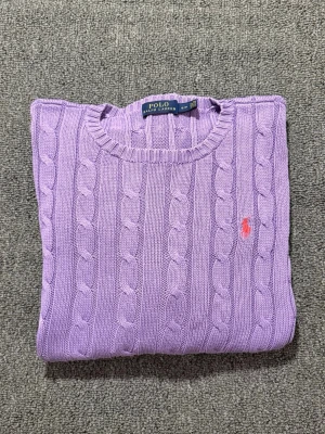 Lila stickad tröja från Polo Ralph Lauren - Säljer en lila kabelstickad tröja från Polo Ralph Lauren i storlek S. Tröjan har rund hals, långärmade ärmar och ett broderat rosa Polo-logo på bröstet. Materialet är mjuk bomull och modellen är klassisk med ribbade muddar vid ärmslut och nederkant.