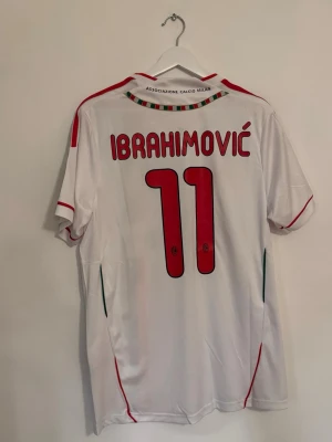 AC Milan Ibrahimović tröja Adidas M - Säljer en officiell AC Milan matchtröja med Ibrahimović och nummer 11 på ryggen. Tröjan är vit med röda och svarta detaljer, Adidas-logga, Fly Emirates tryck och Serie A patch på ärmen. Materialet är polyester och har en klassisk v-ringad krage med röd kant.