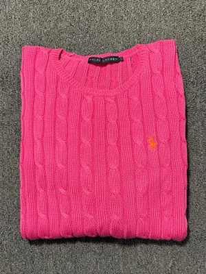 Rosa kabelstickad tröja Ralph Lauren - Säljer en kabelstickad tröja från Ralph Lauren i en stark rosa färg. Tröjan har rund halsringning, långa ärmar och den klassiska broderade loggan i orange på bröstet. Tillverkad i mjuk bomull och perfekt för dig som vill sticka ut lite extra.