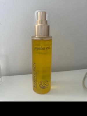 Jojoba Oil från Rosenserien - Jojoba Oil från Rosenserien i en genomskinlig plastflaska med pump och guldfärgad detalj. Oljan är klar och gul, perfekt för återfuktning av huden. Flaskan rymmer 100 ml och har en stilren design med rosdetalj och lock i plast. Nypris: 369kr