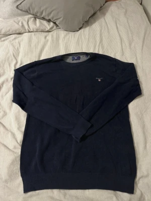 Marinblå stickad tröja från GANT - Snygg marinblå stickad tröja från GANT i storlek L. Tröjan har rund hals, diskret GANT-logga med sköld på bröstet och är tillverkad i premium bomull. Långa ärmar och ribbade muddar vid hals, ärmslut och nederkant. Perfekt för dig som gillar klassisk stil.