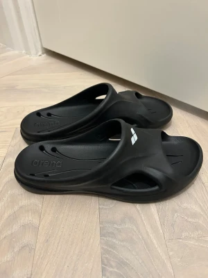 Svarta badsandaler från Arena 37 - Svarta bad sandaler från Arena med sportig design och öppna sidor. Tillverkade i syntetmaterial som torkar snabbt och har en platt sula med mönstrad undersida för extra grepp. Perfekta för poolen eller stranden, med Arena-logga på ovansidan. Bra skick, storlek 37.