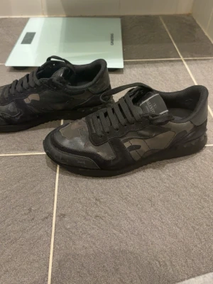 Rockrunners - Säljer ett par svarta Valentino Garavani sneakers med coolt camouflage-mönster. Skorna har snörning, platt sula och rund tå. Materialmix av skinn och mocka ger en lyxig vibe. Perfekta för dig som vill sticka ut med designer på fötterna. Äkta, finns ej box. Köpte för 2400 av en polare när ca 1 år sen