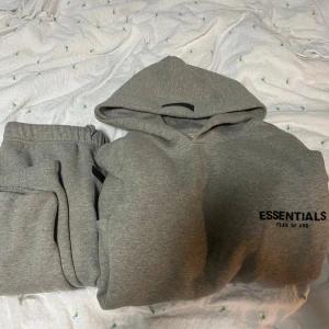 Grå Essentials Fear of God tracksuit - Säljer en grå tracksuit från Essentials Fear of God med svart logga på bröstet och ryggen och mitten av byxorna och nere på byxorna. Tröjan har huva, långa ärmar och är tillverkad i mjuk bomullsmix. Perfekt för dig som gillar streetwear och vill ha en clean look. 
