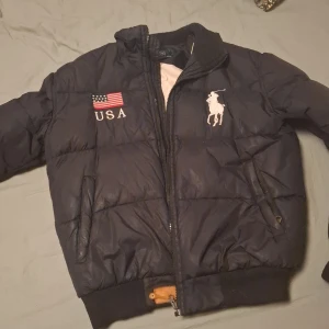 Svart pufferjacka Polo Ralph Lauren - Svart pufferjacka från Polo Ralph Lauren med broderad USA-flagga, stor vit logga och siffran 3 på både fram- och baksida. Jackan har dragkedja, ribbade muddar och är fodrad med dun för extra värme. Perfekt för dig som gillar sportig och amerikansk stil.