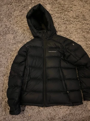 Svart Peak Performance dunjacka - Säljer en svart dunjacka från Peak Performance med huva och dragkedja framtill. Jackan har en puffig look och diskret logga på bröstet samt ärmen.