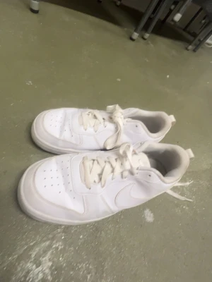 Vita Nike Air Force 1 sneakers - Klassiska helvita Nike Air Force 1 sneakers med lågt skaft och snörning. Skorna har en rund tå, perforerade detaljer på tån och är tillverkade i skinn med en platt sula. Perfekta för en clean och stilren look.