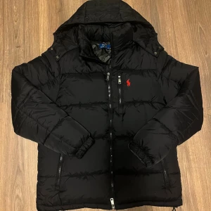 Svart dunjacka från Polo Ralph Lauren - Svart dunjacka från Polo Ralph Lauren med huva, dragkedja och flera fickor. Jackan har quiltad design och en röd Polo-logga på bröstet. Snygga detaljer som snörning vid huvan och RL-knappar. Perfekt för kalla dagar och riktigt stilren look. Med avtagbar luva. Tags medföljer. Fint skicka stl S är för liten på mig som  är ca 185. Justerbar midja. 