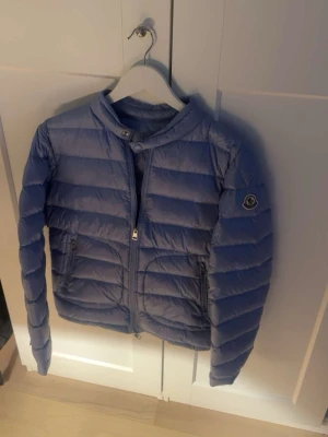 Blå Moncler pufferjacka - Säljer en blå pufferjacka från Moncler, modell Longue Saison. Jackan har ståkrage, dragkedja framtill och två sidofickor med dragkedja. Klassisk Moncler-logga på ärmen. Perfekt för dig som vill ha en lätt och snygg jacka med cool vibe.