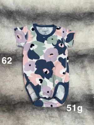 Blommig body i pastellfärger - Supersöt body med kort ärm och blommigt mönster i blått, rosa, lila och grönt. Perfekt för dig som gillar färg och mönster! Bodyn har mjukt material och bekväm passform med tryckknappar i grenen.🌸 Fint använt skick. Storlek 62. 