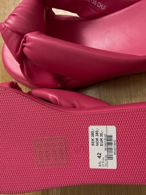 Rosa sandaler med knut från VOX - Säljer ett par chunky rosa sandaler från VOX i storlek 42. De har breda, vadderade band med en stor knutdetalj över foten och är tillverkade i veganskt syntetmaterial. Sulan är platt och bekväm, perfekt för sommarens alla äventyr.