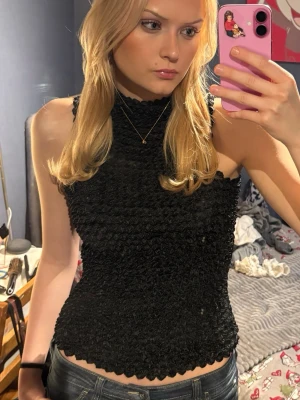 Svart popcorn shirt linne  - Säljer en svart smockad topp med hög hals och ärmlös design. Toppen har en tight passform och är gjord i ett stretchigt syntetmaterial som formar sig snyggt efter kroppen. Perfekt att styla med jeans eller kjol. Popcorn Shirt. 💕