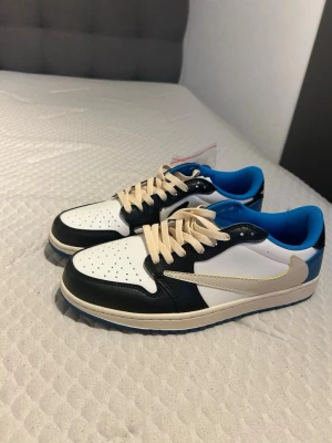 Nike Air Jordan 1 Low Travis Scott x Fragment - Nike Air Jordan 1 Low Travis Scott x Fragment i vit, svart och blå med beige swoosh och snörning. Skorna har broderade detaljer på hälen, blå yttersula och rund tå. Tillverkade i skinn och syntet med ikonisk design och coola kontraster.