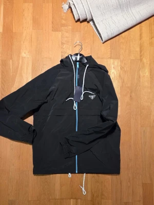 Svart vindjacka från  med blå dragkedja - Säljer en svart vindjacka från Pa med huva och vita snören. Jackan harn snygg blå dragkedja och klassisk Progga på bröstet. Perfekt för dig somill ha en stilren och sportig look. Materialet är lätt och syntetiskt, vilket gör den smidig att bära.