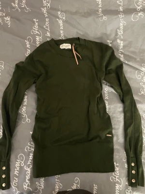 Mörkgrön stickad tröja Holly & White - Snygg mörkgrön stickad tröja från Holly & White med rund hals och långa ärmar. Tröjan har dekorativa knappar vid ärmsluten och en liten metallbricka med logga vid nederkanten. Ribbstickade muddar och en cool randig detalj vid halsen. 💕