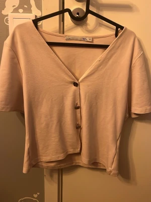 Beige kortärmad topp med guldknappar - Lite mer rosa i verkligheten, Säljer en beige topp från 157 med korta ärmar och v-ringning. Toppen har tre unika, oregelbundna guldknappar framtill som ger en cool touch. Materialet känns mjukt och lätt, perfekt för varmare dagar. Enkel och stilren design som är lätt att matcha.