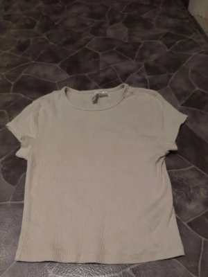 Beige ribbad t-shirt från Divided - Säljer en beige ribbad t-shirt från Divided med rund hals och korta ärmar. T-shirten är enkel och stilren, perfekt för att matcha med jeans eller kjol. Materialet är mjukt och skönt bomull som sitter bekvämt mot kroppen.