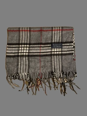 Grå rutig halsduk från Burberrys - Säljer en klassisk grå halsduk från Burberrys med rutigt mönster i svart, vitt och rött. Halsduken är tillverkad i 100% lammull och har fransar i ändarna. Perfekt accessoar för att lyfta din outfit. Perfekt nu när snön har kommit❄️ Kom med frågor och prisförslag!