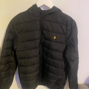 Svart pufferjacka från Lyle & Scott - Säljer en svart pufferjacka från Lyle & Scott med huva och gul logga på. Jackan är i bra skick.