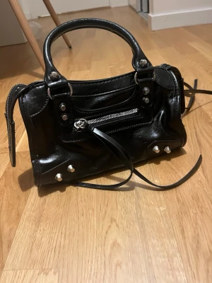Svart handväska med nitar och dragkedja -  Svart snygg handväska i skinn med silverfärgade nitar och detaljer. Väskan är i mycket bra skick. Den har en dragkedja upptill och en extra dragkedja framtill. Den har snygga långa remmar och insidan är rymlig med två fack. Köptes för 250kr. Funderingar? Skriv till mig💕