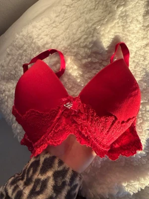 Röd spetsdetaljerad BH U Bella C85 -  röd BH från U Bella med vadderade kupor och spetsdetaljer längs kanterna. Dekorerad med en liten rosett och metallcharm framtill. Justerbara axelband och mjukt material som ger en bekväm passform. Köpt i fel storlek så har aldrig kommit till användning tyvärr!