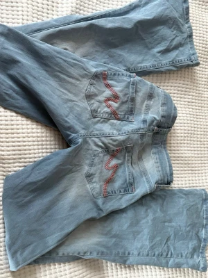 Bootcut ljusblå jeans från Nelly - Säljer ett par ljusblå bootcut jeans från Nelly med breda ben och klassisk femficksdesign. Baksidan har snygga fickor med rödprickig broderad detalj. Jeansen har dubbla knappar i midjan och är tillverkade i mjukt denimtyg.