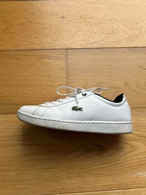 Vita sneakers från Lacoste - Klassiska vita sneakers från Lacoste med den ikoniska gröna krokodilen på sidan. Skorna har rund tå, snörning och svart detalj vid hälen. Ovandelen är i skinn och sulan är platt och mönstrad för bra grepp. Perfekta till jeans eller shorts.