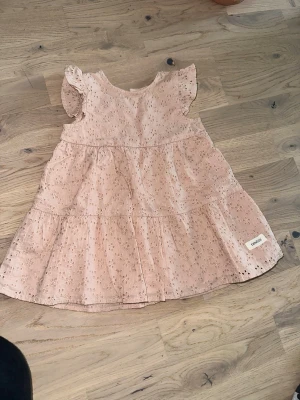 Ljusrosa klänning från Newbie Newbie rosa klänning, storlek 74 - Söt ljusrosa kortklänning från Newbie med broderade blommor och volangärmar. Klänningen har knappar baktill och är gjord i mjuk bomull. Perfekt för varma dagar och har en romantisk känsla med sina fina detaljer. Söt, ljusrosa klänning med broderade detaljer och volangärmar. Klänningen har en trelagerskjol och knäpps med knappar i ryggen.