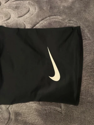 Svart Nike Dri-Fit träningshalsduk - Svart träningshalsduk från Nike med Dri-Fit-teknologi som håller dig torr och bekväm. Västen har en stilren design med ett vitt Nike Swoosh-tryck framtill. Materialet är syntet och passar perfekt för sport och aktivitet.