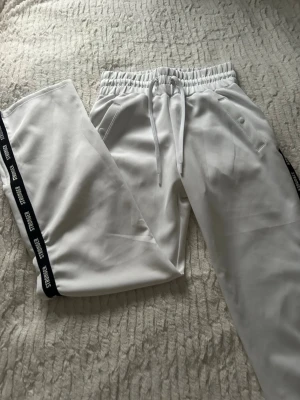 Vita byxor  från Stronger - Snygga vita byxor  från Stronger med svarta sidoremsor där det står 'STRONGER' i vitt. Byxorna har elastisk midja med snörning och praktiska fickor med knapp. Materialet känns mjukt och skönt, perfekt för chill eller träning.