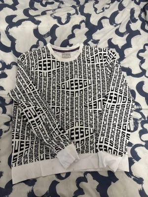 Vit och svart sweatshirt från Guess - Säljer en vit sweatshirt från Guess med svart grafiskt tryck över hela tröjan där det står 'GUESS' i ett coolt mönster. Tröjan har rund hals, långa ärmar och ribbade muddar vid ärmslut och nederkant. Perfekt för dig som gillar streetwear och vill sticka ut.