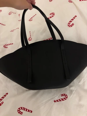 Viral handväska från Gina tricot - Stilren svart handväska från Gina med unik, bred form och långa handtag. Väskan är tillverkad i syntetmaterial och har en enkel, minimalistisk design utan mönster. Insidan har en praktisk krok för nycklar eller accessoarer. Perfekt för dig som gillar clean och modern look. Oanvänd och prislapp kvar!