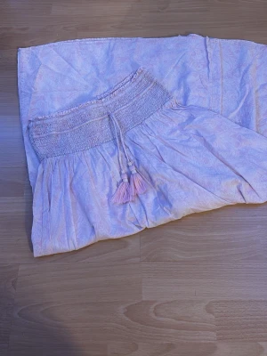 Ljusrosa långkjol från Gina Tricot - Supersöt ljusross långkjol från Gina Tricot med smock i midjan och dekorativa tofsar. Kjolen har ett diskret paisleymönster i pastelliga toner och är gjord i ett tunt, flowy material som känns luftigt och skönt. Perfekt för dig som gillar boho-vibbar och mjuka färger.