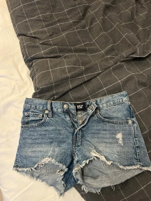 Blå jeansshorts från 157, XS - Säljer ett par blå jeansshorts från 157 i storlek XS. De har råa, fransiga kanter och slitna detaljer för en avslappnad look. Klassisk femficksmodell med knapp och dragkedja framtill. Perfekta till sommaren!