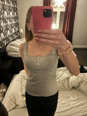 Grå ribbad linnetopp med knappar - Säljer en ljusgrå ribbad linnetopp i storlek S. Toppen har rundad halsringning och tre snygga pärlemorknappar framtill. Perfekt till jeans eller shorts och superenkel att styla. Materialet är mjukt och stretchigt för en bekväm passform.