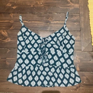 Blå linnetopp med vita blad - Säljer en blå linnetopp med vita bladmönster och snörning framtill. Toppen har smala axelband och en lätt utsvängd nederkant. Perfekt för varma dagar och enkel att matcha med jeans eller shorts.
