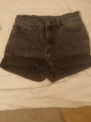 Svarta denimshorts från UNIQ YOUNG - Säljer ett par svarta jeansshorts från UNIQ YOUNG i storlek 146. De har hög midja, klassiska fem fickor och uppvikta benslut. Materialet är slitstark bomullsdenim med snygga sömmar och knappstängning framtill. Perfekt för dig som gillar enkel och cool stil.