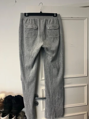 Grå mjukisbyxor Juicy Couture M - Säljer ett par grå mjukisbyxor från Juicy Couture i storlek M. Byxorna har snörning i midjan med silverfärgade detaljer, raka ben och fickor både fram och bak med lock och knapp. Materialet är mjukt och skönt, perfekt för chill dagar.