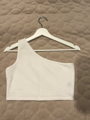 Vit oneshoulder croppad topp - Säljer en vit ribbad oneshoulder topp som är croppad och har en snygg asymmetrisk design. Perfekt för dig som gillar minimalistisk stil och vill ha något som sticker ut lite. Materialet är mjukt och stretchigt, vilket gör den bekväm att bära.