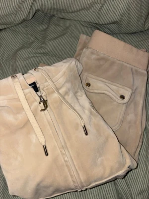 Juicy Couture XS - Säljer ett par beige mjukisbyxor från Juicy Couture i storlek XS. Byxorna har snörning framtill. Materialet är mjukt och sammet-liknande, med fickor och detaljer i ändarna av snörerna och på fickorna 