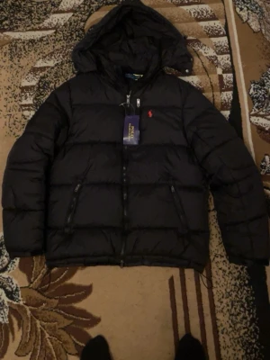 Svart pufferjacka Polo Ralph Lauren - Säljer en svart pufferjacka från Polo Ralph Lauren i storlek M. Jackan har huva, två sidofickor med dragkedja och den klassiska röda loggan på bröstet. Materialet är syntet och jackan har en quiltad look med dubbla dragkedjor framtill.