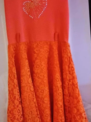 Orange spetsklänning med hjärtdetalj - Säljer en orange kortklänning med spetskjol och ett glittrigt hjärta av stenar på bröstet. Överdelen är slät medan kjolen har ett blommigt spetsmönster. Perfekt för dig som vill sticka ut med färg och detaljer.