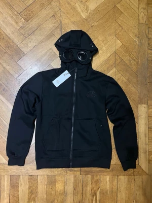 Cp company zip hoodie - Svart Cp company zip hoodie med luva. Finns ett litet hack på högra ögat. Helt ny!