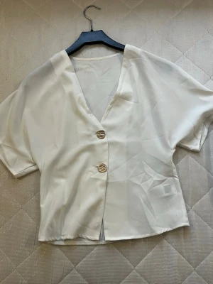 Vit blus med v-ringning och knappar - Vero Moda  - Säljer en stilren vit blus med korta ärmar och v-ringad hals. Blusen har två dekorativa guldfärgade knappar framtill och är tillverkad i ett mjukt, lätt material som ger en skön känsla. Perfekt för dig som gillar enkel och clean stil.