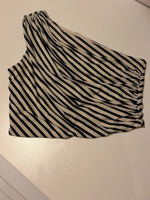Randig oneshoulder topp i svart och vitt! - Säljer randig topp med oneshoulder-design i svart och vitt som tyvärr inte passar mig! Skönt svalt tyg, perfekt till sommaren då den är lite kortare i modellen! Köpt secondhand men är som ny! Ser gul ut på bilden men är mer krämvit! Storlekslappen är bort så svårt att säga men ungefär xs/s!