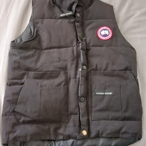 Canada goose väst - Svart dunväst från Canada Goose med klassisk logotyp på bröstet. Västen har två stora fickor framtill, knappar och dragkedja. Materialet är polyester och dun, perfekt för kalla dagar. Snygg och stilren design med hög krage och quiltad yta. HAG HAR DÅLIG MOBIL SÅ DE BLIR DÅLIGA BILDER