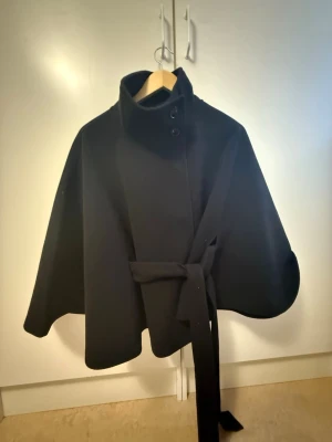 Svart kappa med bälte från Gina Tricot - Svart kappa från Gina Tricot i storlek XS/S. Snygg cape-modell med hög krage, knappar framtill och ett matchande bälte i midjan. Materialet är mjukt och känns som ull. Perfekt för dig som vill ha en stilren och cool look.